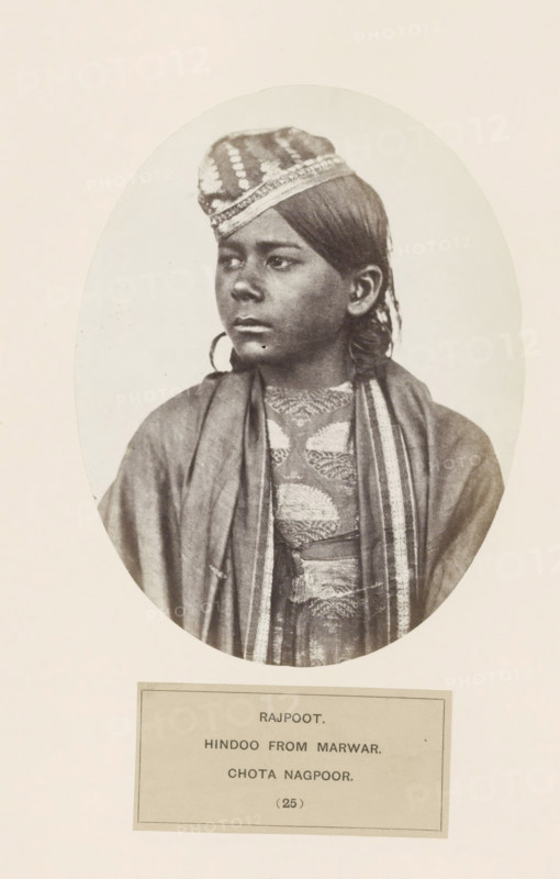 Portrait of an unknown man from the Rajput community originally from Jodhpur in Rajasthan, in Chota Nagpur in eastern India, photograph, page, number: '(25)', inscription: 'RAJPOOT., HINDOO FROM MARWAR., CHOTA NAGPOOR.', photographer: Benjamin Simpson (possibly), Chota Nagpur, c. 1858 - in or before 1868, Jharkhand, paper, albumen print, height 158 mm x width 122 mm, human races, peoples, nationalities, head-gear: cap, A sepia-toned oval portrait shows a young person with dark hair pulled back and wearing a patterned cap tilted slightly forward on the head, looking to the side with a neutral expression; they wear layered garments including a shawl draped over the shoulders and a textured, patterned garment beneath with visible trim along the edges, and the portrait is mounted on a light background with a rectangular label area below.