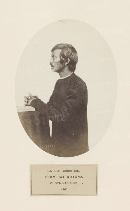 A praying Christian man from Rajput community in a kneeling position in Chota Nagpur, eastern India, photograph, page, inscription: 'RAJPOOT CHRISTIAN., FROM RAJPOOTANA., CHOTA NAGPOOR.', number: '(26)', photographer: Benjamin Simpson (possibly), Chota Nagpur, c. 1858 - in or before 1868, Jharkhand, paper, albumen print, height 175 mm x width 129 mm, one person praying, human races, peoples, nationalities, A sepia-toned oval photograph shows a young man in profile facing left, with short dark hair and a mustache, wearing a dark long-sleeved garment with a lighter collar visible at the neck; he is posed with his hands clasped and resting on a flat surface, and the oval image is set against a pale rectangular background with a separate rectangular label area below the portrait.