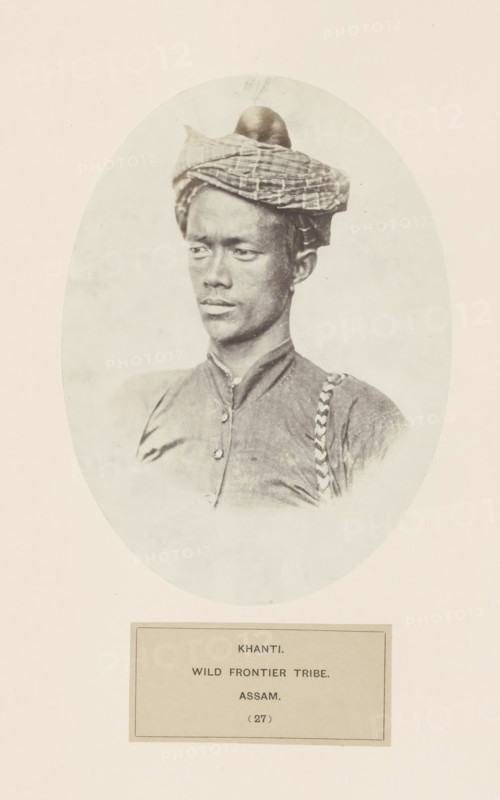 Portrait of an unknown man from the Khamti tribe and Arunachal Pradesh, photograph, page, inscription: 'KHANTI., WILD FRONTIER TRIBE., ASSAM.', number: '(27)', photographer: attributed to Benjamin Simpson, Assam, c. 1858 - in or before 1868, Attributed to, paper, albumen print, height 176 mm x width 129 mm, head-gear: turban, human races, peoples, nationalities, Arunachal Pradesh, A sepia-toned oval portrait of a young man shown from the chest up, wearing a patterned cloth wrapped around his head and a buttoned shirt with a braided strap over one shoulder; he has a neutral expression and looks slightly to his left, with a plain light background surrounding him and a rectangular plaque positioned below the oval.