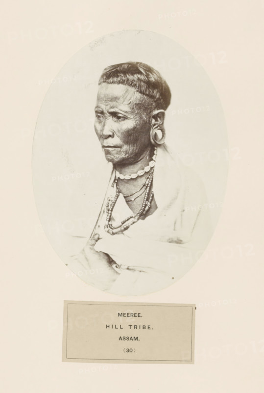 Portrait of an unknown man from the Miri tribe of Assam, photograph, page, The Miri tribe is also known as Mising., inscription: 'MEEREE., HILL TRIBE., ASSAM.', number: '(30)', photographer: attributed to Benjamin Simpson, Assam, c. 1858 - in or before 1868, Attributed to, paper, albumen print, height 175 mm x width 130 mm, human races, peoples, nationalities, An oval-framed black-and-white portrait of an older person shown from the chest upward, facing slightly left with a neutral expression and visible facial wrinkles, a short straight fringe haircut, large circular ear ornaments, multiple layered necklaces including a strand of shell-like beads, a draped light-colored cloth over the shoulders, and one hand holding the cloth near the chest; the portrait sits on a pale background with a rectangular label area below.