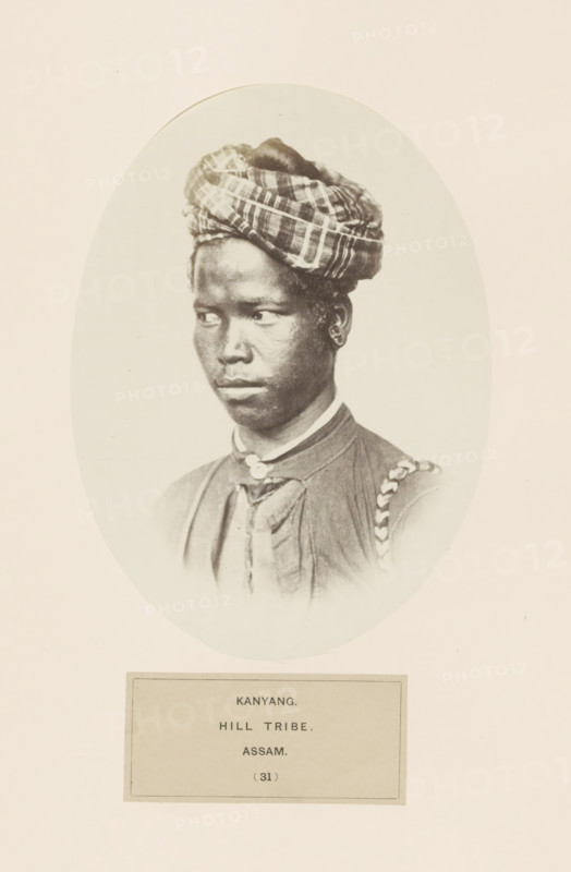 Portrait of an unknown man from the Ahom community of Assam, photograph, page, The larger ethnic group is known as Shan in Myanmar., number: '(31)', inscription: 'KANYANG., HILL TRIBE., ASSAM.', photographer: Benjamin Simpson (possibly), Assam, c. 1858 - in or before 1868, paper, albumen print, height 177 mm x width 129 mm, human races, peoples, nationalities, A sepia-toned oval portrait of a person with dark skin wearing a patterned headwrap and a high-collared garment with visible buttons and a braided strap over one shoulder, shown from the chest up against a plain light background, with a rectangular plaque beneath the oval.