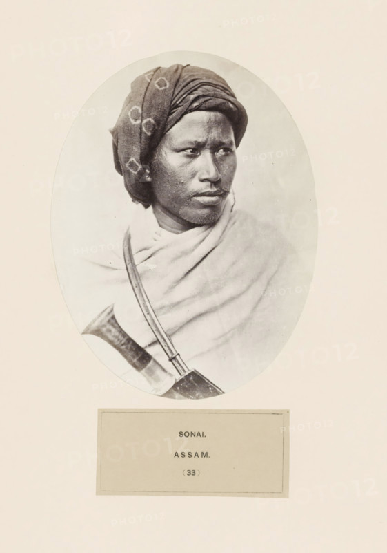 Portrait of an unknown man from the region of Sonai in Assam, photograph, page, number: '(33)', inscription: 'SONAI., ASSAM.', photographer: Benjamin Simpson (possibly), Assam, c. 1858 - in or before 1868, paper, albumen print, height 157 mm x width 120 mm, human races, peoples, nationalities, A sepia-toned oval portrait of a person shown from the shoulders up wearing a patterned headwrap and a draped cloth over the shoulders, the face turned slightly to one side with a neutral expression, visible facial hair and textured skin, and a curved object with a strap crossing the chest partially visible at the lower edge.