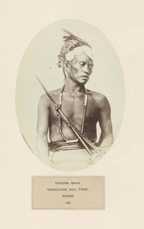 Portrait of an unknown man from the Rengma Naga tribe of Nagaland and Assam, photograph, page, number: '(36)', inscription: 'RENGMA NAGA., MARAUDING HILL TRIBE., ASSAM', photographer: Benjamin Simpson (possibly), Assam, c. 1858 - in or before 1868, paper, albumen print, height 177 mm x width 129 mm, human races, peoples, nationalities, casting weapons: spear, Nagaland, A sepia-toned oval photograph shows a bare-chested man seated and holding a long spear diagonally across his torso, wearing a headpiece with decorative elements and circular adornments at his temples, elongated earrings, and multiple beaded necklaces; a cloth is wrapped around his waist and his gaze is turned to the side.