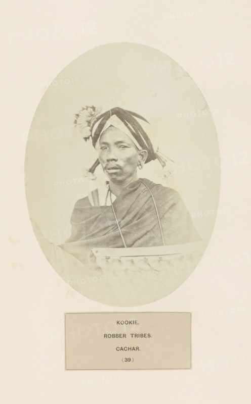 Portrait of an unknown man from the Kuki tribe of Assam, photograph, page, number: '(39)', inscription: 'KOOKIE., ROBBER TRIBES., CACHAR.', photographer: Benjamin Simpson (possibly), Cachar, c. 1858 - in or before 1868, Assam, paper, albumen print, height 175 mm x width 129 mm, human races, peoples, nationalities, A faded oval portrait of a person wearing a patterned headwrap with fabric tassels and a decorative floral adornment, visible earrings, and a draped garment; the person is seated behind a carved wooden or woven object with a scalloped edge, and the portrait is mounted on a pale background with a rectangular label panel below.