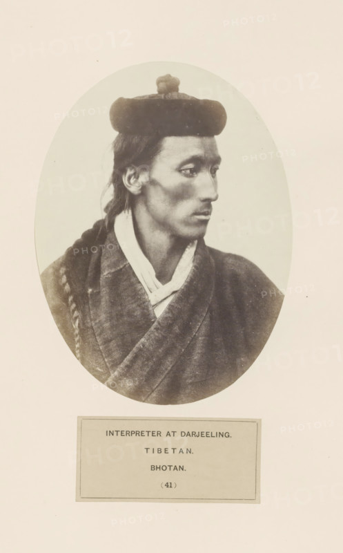Portrait of an unknown man from Bhutan in Darjeeling, West Bengal, photograph, page, number: '(41)', inscription: 'INTERPRETER AT DARJEELING., TIBETAN., BHOTAN.', photographer: Benjamin Simpson (possibly), Darjiling, c. 1858 - in or before 1868, Bengal, Northeast India, paper, albumen print, height 160 mm x width 120 mm, head-gear: hat, human races, peoples, nationalities, A sepia photographic portrait shows a person in an oval frame facing right with a composed expression and a narrow face. The person wears a rounded hat with a small rounded adornment on top and has long hair pulled back, one section braided and resting over a shoulder. They are dressed in layered garments with a visible collar and a textured outer robe that crosses at the front. The background inside the oval is plain and light, and the oval image is set on a larger pale background with a rectangular label area below.
