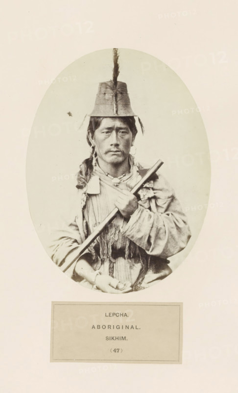 Portrait of an unknown man with a weapon from the Lepcha community of Sikkim, photograph, page, number: '(47)', inscription: 'LEPCHA., ABORIGINAL., SIKHIM.', photographer: Benjamin Simpson (possibly), Sikkim, c. 1858 - in or before 1868, paper, albumen print, height 159 mm x width 120 mm, head-gear: hat, human races, peoples, nationalities, casting weapons, A sepia-toned oval portrait of a person sitting and facing the camera, wearing a tall hat with a vertical ornament extending upward, earrings, and layered clothing with fringed or textured fabric; the person holds a long cylindrical object diagonally across their chest with one hand near the top and the other resting lower, and their expression is neutral.