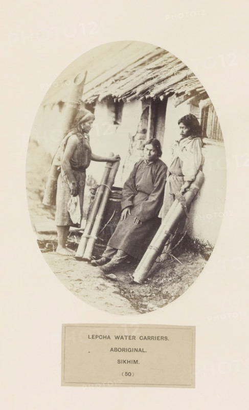 Three water carriers in front of a house from the Lepcha community of Sikkim, photograph, page, number: '(50)', inscription: 'LEPCHA WATER CARRIERS., ABORIGINAL., SIKHIM.', photographer: Benjamin Simpson (possibly), Sikkim, c. 1858 - in or before 1868, paper, albumen print, height 176 mm x width 129 mm, street, human races, peoples, nationalities, An oval sepia photograph shows three people posed in front of a thatched building; one person sits on a low ledge wearing a long dark garment and boots with hands resting on their knees, another stands to the left holding a tall cylindrical bundle and wearing a sleeveless top and a skirt with bare feet, and the third stands to the right leaning against the wall with a wrapped bundle by their side and wearing a long-sleeved shirt and skirt; large cylindrical containers made of a smooth material are propped against the wall and ground, the building has a rough plastered exterior and a sloping thatch roof with visible straw edges, and the ground...