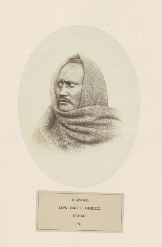 Portrait of an unknown man from the Rajwar community of Bihar in eastern India, photograph, page, number: '(7)', inscription: 'RUJWAR., LOW CASTE HINDOO., BEHAR.', Bihar, c. 1858 - in or before 1868, photographer, North India, paper, albumen print, height 160 mm x width 120 mm, human races, peoples, nationalities, A sepia-toned oval portrait of a person wrapped in a textured shawl that covers the head and shoulders, showing a close-up three-quarter profile with a serious facial expression, short hair visible at the forehead, prominent cheekbones, a mustache, and deep-set eyes looking toward the left, against a plain light background and surrounded by a wide blank border.