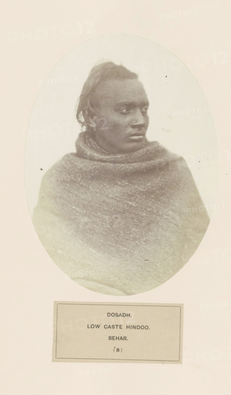 Portrait of an unknown man from the Dusadh community of Bihar in eastern India, photograph, page, The Dusadh community is also known as Paswan., number: '(8)', inscription: 'DOSADH., LOW CASTE HINDOO., BEHAR.', Bihar, c. 1858 - in or before 1868, photographer, North India, paper, albumen print, height 175 mm x width 129 mm, human races, peoples, nationalities, An oval-framed, sepia-toned portrait of a person with dark skin shown from the chest upward, facing slightly to the viewer's right, with short hair pulled back and a serious facial expression; the person is wrapped in a textured, heavy shawl or blanket that covers the shoulders and chest, and the portrait is mounted on a pale rectangular background with a bordered label area below the oval.