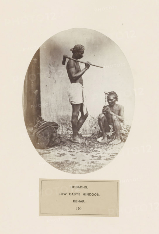 Two unknown men with tools from the Dusadh community of Bihar in eastern India, photograph, page, The Dusadh community is also known as Paswan., inscription: 'DOSADHS., LOW CASTE HINDOOS., BEHAR.', number: '(9)', Bihar, c. 1858 - in or before 1868, photographer, North India, paper, albumen print, height 158 mm x width 121 mm, human races, peoples, nationalities, A sepia-toned oval photograph shows two barefoot men against a plain wall. One man stands with a cloth wrapped around his waist and a headscarf, holding a long-handled tool resting on his shoulder and looking downward. The other man sits on the ground with a cloth around his lower body, holding a small object vertically in both hands and looking at it. There are cloth bundles and scattered debris on the ground near their feet.