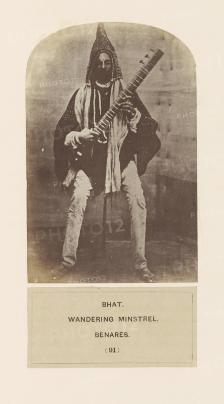 Portrait of an unknown minstrel (Bhat) from Varanasi, Uttar Pradesh, India, photograph, page, inscription, q: 'BHAT., WANDERING MINSTREL., BENARES.', number: '(91)', Varanasi, c. 1858 - in or before 1868, photographer, North India, paper, albumen print, height 134 mm x width 88 mm, minstrel, 'juggler', human races, peoples, nationalities, A monochrome photograph of a seated person wearing a tall conical hat and layered loose clothing, holding a long-necked stringed instrument across their body; the person has a beard and is draped with scarves or cloths, and their hands rest on the instrument’s neck and body. The figure sits against a plain backdrop within an arched frame and wears light-colored trousers and dark footwear. Below the photograph is a rectangular panel containing blocks of printed lines.