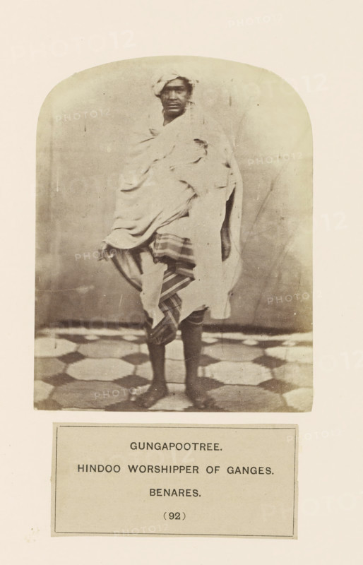 Portrait of an unknown man from the Gangaputra Brahmin community of Varanasi, Uttar Pradesh, India, photograph, page, inscription: 'GUNGAPOOTREE., HINDOO WORSHIPPER OF GANGES., BENARES.', number: '(92)', Varanasi, c. 1858 - in or before 1868, photographer, North India, paper, albumen print, height 122 mm x width 95 mm, Hinduism, Buddhism, Jainism, human races, peoples, nationalities, A full-length sepia photograph of a person standing on a patterned floor, wrapped in layered light-colored drapery with a striped cloth visible around the lower body, a cloth wrapped around the head, bare legs and feet showing, and a plain backdrop with rounded top framing the image.