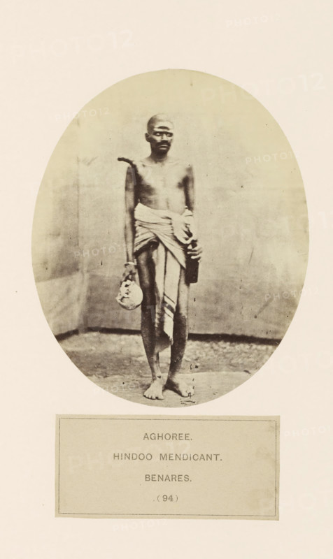 Portrait of an unknown man from the Aghori mendicant community of Varanasi, Uttar Pradesh, India, photograph, page, inscription: 'AGHOREE., HINDOO MENDICANT., BENARES.', number: '(94)', Varanasi, c. 1858 - in or before 1868, photographer, North India, paper, albumen print, height 128 mm x width 105 mm, Hinduism, Buddhism, Jainism, human races, peoples, nationalities, A sepia-toned oval photograph of a barefoot man standing against a plain backdrop, his torso bare and a cloth wrapped around his waist and draped over one shoulder, holding a small round object in one hand and a rectangular object in the other, with a neutral facial expression and a shaved head, the image set on a light-colored page with a rectangular bordered area below the oval.