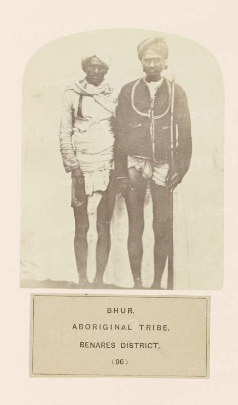 Portrait of two unknown men from the Bhurtiya community of Varanasi district, Uttar Pradesh, India, photograph, page, number: '(96)', inscription: 'BHUR., ABORIGINAL TRIBE., BENARES DISTRICT.', Varanasi, c. 1858 - in or before 1868, photographer, North India, paper, albumen print, height 123 mm x width 96 mm, human races, peoples, nationalities, A faded sepia photograph framed by a rounded top shows two standing men wearing turbans and wrapped cloth garments, both barefoot and facing forward; the man on the right wears a darker jacket-like top with a looped cord over his shoulders and holds a long staff, while the man on the left wears lighter layered wraps around his torso and head and has one arm hanging by his side.