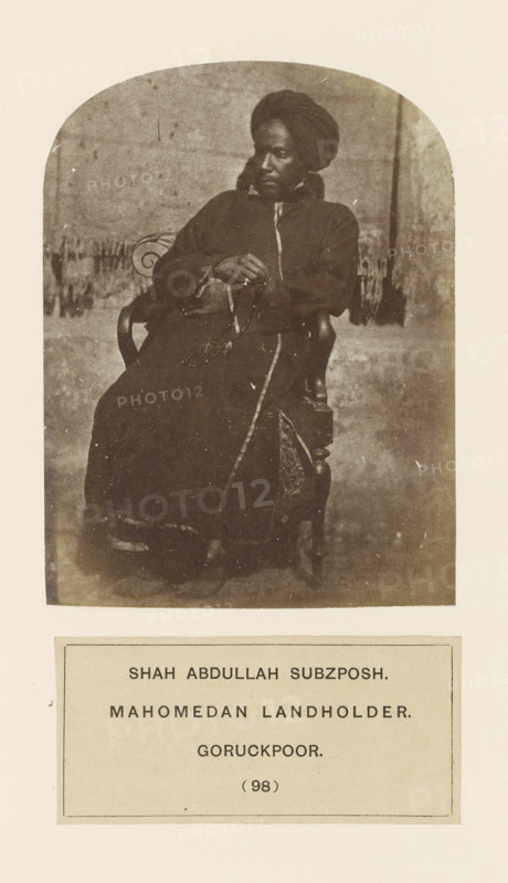 Portrait of Shah Abdullah Subzposh from Gorakhpur, Uttar Pradesh, India, photograph, page, number: '(98)', inscription: 'SHAH ABDULLAH SUBZPOSH., MAHOMEDAN LANDHOLDER., GORUCKPOOR.', Gorakhpur, c. 1858 - in or before 1868, photographer, North India, paper, albumen print, height 117 mm x width 88 mm, human races, peoples, nationalities, A sepia-toned photograph shows a seated person wearing a dark, full-length garment and a wrapped head covering, leaning slightly back in a wooden chair with ornate armrests; the person's hands rest in their lap holding a string of beads and a thin stick, and a patterned fabric panel is visible on the chair beside them, while a plain backdrop and a fringed textile behind the chair are seen in the upper portion of the image and a blank margin surrounds the photograph.