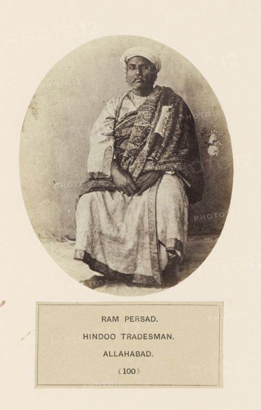 Portrait of a tradesman Ram Persad from Prayagraj, Uttar Pradesh, India, photograph, page, Prayagraj is also known as Allahabad., inscription: 'RAM PERSAD., HINDOO TRADESMAN., ALLAHABAD.', number: '(100)', Allahabad, c. 1858 - in or before 1868, photographer, North India, paper, albumen print, height 119 mm x width 97 mm, head-gear: turban, human races, peoples, nationalities, persons engaged in trade and commerce, A sepia-toned oval photograph shows a seated man wearing a turban and traditional layered garments, including a patterned draped cloth over one shoulder and a lighter robe underneath; his hands rest together in his lap and his feet are visible beneath the long robe, with a plain background surrounding him and a rectangular label area positioned below the oval.