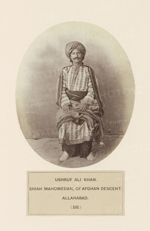 Portrait of Ushruf Ali Khan from Prayagraj, Uttar Pradesh, India, photograph, page, Prayagraj is also known as Allahabad., number: '(101)', inscription: 'USHRUF ALI KHAN., SHIAH MAHOMEDAN, OF AFGHAN DESCENT., ALLAHABAD.', Allahabad, c. 1858 - in or before 1868, photographer, North India, paper, albumen print, height 129 mm x width 104 mm, head-gear: turban, human races, peoples, nationalities, An oval sepia photograph of a seated man wearing a wrapped turban and patterned garments with layered fabric draped over his shoulders and lap, his hands resting on the cloth in front of him, visible facial hair and a neutral expression, bare feet peeking from beneath his clothing, all set against a plain backdrop with a rectangular framed area below the oval photograph.