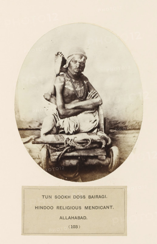 Portrait of Tun Sookh Doss, a mendicant from Prayagraj, Uttar Pradesh, India, photograph, page, Prayagraj is also known as Allahabad., inscription: 'TUN SOOKH DOSS BAIRAGI., HINDOO RELIGIOUS MENDICANT., ALLAHABAD.', number: '(103)', Allahabad, c. 1858 - in or before 1868, photographer, North India, paper, albumen print, height 128 mm x width 105 mm, monks and nuns, human races, peoples, nationalities, A sepia-toned oval photograph showing a seated man with a cloth wrapped around his head and draped garments, beads around his neck, arms crossed over his chest, and one foot visible behind his shoulder; he sits on a rolled bundle and a small platform with ropes, set against a plain backdrop, framed by a light margin.