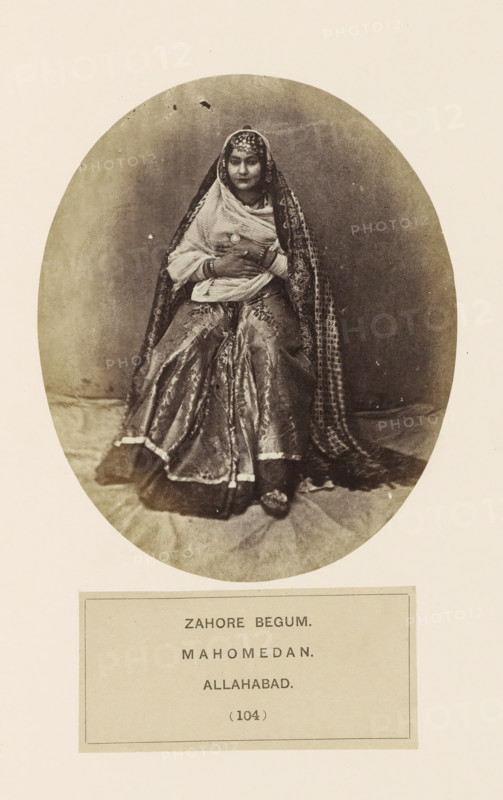 Portrait of Zahore Begum, a courtesan from Prayagraj, Uttar Pradesh, India, photograph, page, Prayagraj is also known as Allahabad., number: '(104)', inscription: 'ZAHORE BEGUM., MAHOMEDAN., ALLAHABAD.', Allahabad, c. 1858 - in or before 1868, photographer, North India, paper, albumen print, height 128 mm x width 105 mm, human races, peoples, nationalities, courtesan, hetaera, A sepia-toned oval photograph shows a seated woman wearing layered traditional garments, including a patterned outer drape that covers her head and shoulders and a richly textured skirt that spreads around her. Her hands are folded at her midsection and she wears bracelets and a circular brooch. The head covering includes decorative detailing across the forehead and the drape falls in patterned folds beside her. The background inside the oval is plain and slightly mottled, and the lower area shows a fabric surface beneath her feet. The photograph is mounted on a lighter rectangular page with a bordered panel belo...