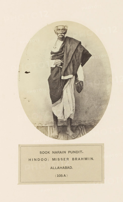 Portrait of Sook Narain, a high priest from Prayagraj, Uttar Pradesh, India, photograph, page, Prayagraj is also known as Allahabad., number: '(105 A )', inscription: 'SOOK NARAIN PUNDIT., HINDOO: MISSER BRAHMIN., ALLAHABAD.', Allahabad, c. 1858 - in or before 1868, photographer, North India, paper, albumen print, height 120 mm x width 95 mm, human races, peoples, nationalities, priest, Hinduism, Buddhism, Jainism, An oval sepia photograph shows an older man standing on a narrow wooden floor, wearing a wrapped garment draped over one shoulder and a light cloth around his lower body, with a cloth cap on his head and a shawl folded across his arm, his feet are bare and his hands are held near his midsection, and the oval image is set against a light rectangular background with a bordered panel beneath it.