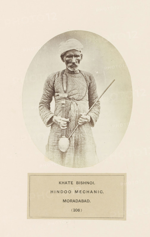 Portrait of Khate with a hookah, a man from the Bishnoi community of Moradabad, Uttar Pradesh, India, photograph, page, inscription: 'KHATE BISHNOI., HINDOO MECHANIC., MORADABAD.', number: '(106)', Moradabad, c. 1858 - in or before 1868, photographer, North India, paper, albumen print, height 120 mm x width 95 mm, water-pipe, hookah, narghile, human races, peoples, nationalities, An oval-framed sepia photograph of an older man standing and facing forward, wearing a wrapped head cloth and a patterned long garment, holding a long thin rod in one hand and a tool with a long handle and rounded end in the other, with a rectangular label area below the oval.