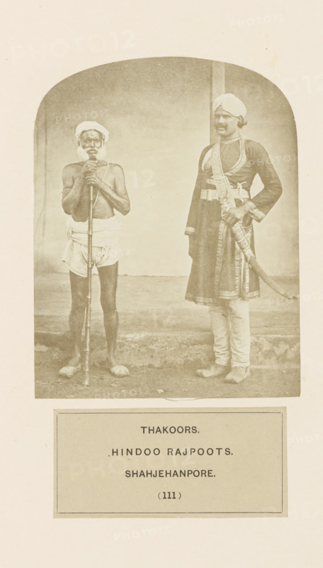 Portrait of two unknown men from the Thakur community of Shahjahanpur, Uttar Pradesh, India, photograph, page, number: '(111)', inscription: 'THAKOORS., HINDOO RAJPOOTS., SHAHJEHANPORE.', Shahjahanpur, c. 1858 - in or before 1868, photographer, North India, paper, albumen print, height 124 mm x width 96 mm, human races, peoples, nationalities, A sepia photograph shows two men standing side by side against a plain background with a curved top border. The man on the left is shirtless and wearing a simple cloth wrapped around his waist and a turban on his head, holding a long staff vertically in front of him with both hands and standing barefoot. The man on the right is dressed in a long ornate coat with patterned trousers and a turban, wearing decorative belts and a sash, with one hand resting on a sheathed curved sword at his side and his feet positioned slightly apart. The ground beneath them appears worn and the image is framed by a light border with a rectangular panel below the phot...