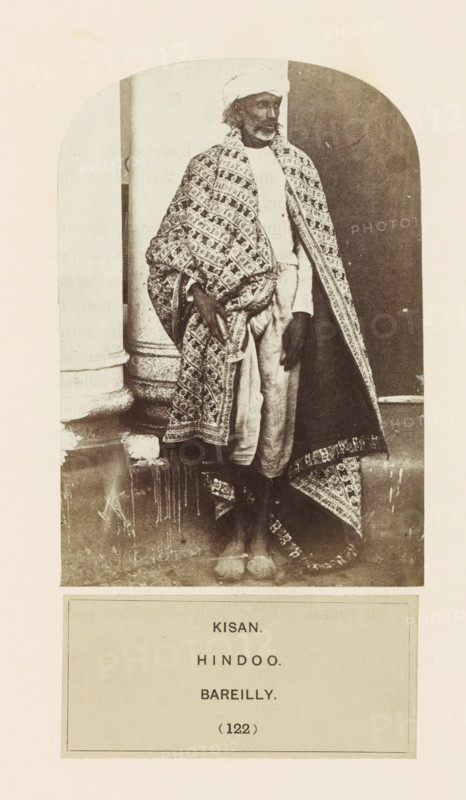 Portrait of an unknown man from the Kisan community of Bareilly, Uttar Pradesh, India, photograph, page, inscription: 'KISAN., HINDOO., BAREILLY.', number: '(122)', Bareilly, c. 1858 - in or before 1868, photographer, North India, paper, albumen print, height 127 mm x width 88 mm, human races, peoples, nationalities, A sepia photograph shows a standing man wearing a patterned shawl draped over his shoulders and wrapped around his body, a light turban on his head, a light long-sleeved shirt, and loose light trousers; he has a beard and mustache, his hands rest near his hips, and he stands barefoot on a step in front of a column and a dark doorway, with the edge of the patterned shawl trailing to the ground.