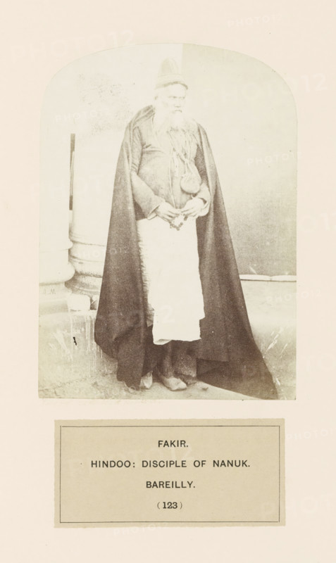 Portrait of a fakir (ascetic) from the Nanakshahi community of Bareilly, Uttar Pradesh, India, photograph, page, inscription: 'FAKIR., HINDOO: DISCIPLE OF NANUK., BAREILLY.', number: '(123)', Bareilly, c. 1858 - in or before 1868, photographer, North India, paper, albumen print, height 133 mm x width 96 mm, human races, peoples, nationalities, asceticism, Hinduism, Buddhism, Jainism, A sepia-toned photograph shows an older man standing inside an arched frame, wearing a tall pointed cap, a long dark cloak that drapes to the floor, a light apron-like cloth over his lower body, and layered garments beneath; he has a long beard and holds a string of beads or small objects in his hands while a column and a plain backdrop are visible behind him.