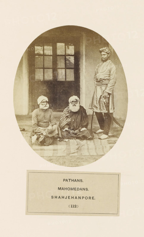 Three unknown men from the Pathan community of Shahjahanpur, Uttar Pradesh, India, photograph, page, inscription: 'PATHANS.., MAHOMEDANS., SHAHJEHANPORE.', number: '(112)', Shahjahanpur, c. 1858 - in or before 1868, photographer, North India, paper, albumen print, height 129 mm x width 104 mm, human races, peoples, nationalities, An oval sepia photograph shows three men in front of a building entrance with a paneled door behind them; two men are seated on a patterned cloth on the ground, both wearing turbans and long garments with one having a long white beard and a draped shawl, and the third man stands to their side wearing a patterned tunic, a wrapped turban, and holding a curved sword with scabbard while wearing light-colored footwear.