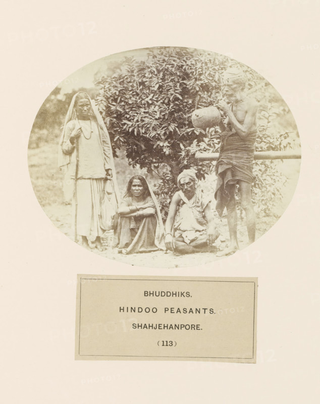 Four unknown Bhuddhiks from Shahjahanpur in front of a bush, Bhuddhiks. Hindu peasants. Shahjehanpore, photograph, page, number: '(113)', Shahjahanpur, c. 1858 - in or before 1868, photographer, North India, Uttar Pradesh, paper, albumen print, height 104 mm x width 129 mm, human races, peoples, nationalities, A faded circular photograph shows four people positioned in front of a leafy bush, with two individuals seated on the ground and two standing. On the left a person stands wearing a long light garment with a cloth draped over the head and shoulders, holding a staff and a bag. In the center foreground two seated figures face the camera; one has a cloth covering the head and rests arms on folded knees, the other wears a wrapped head covering and a draped upper garment with hands resting on the ground. On the right a thin standing figure wears a wrapped lower cloth and a head covering while holding a rounded pot near the face. The background includes foliage and a horizontal wooden r...