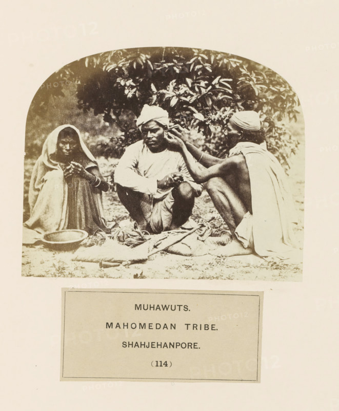Three unknown people from the Muhawut community of Shahjahanpur, Uttar Pradesh, India, photograph, page, inscription: 'MUHAWUTS., MAHOMEDAN TRIBE., SHAHJEHANPORE.', number: '(114)', Shahjahanpur, c. 1858 - in or before 1868, photographer, North India, paper, albumen print, height 98 mm x width 118 mm, human races, peoples, nationalities, A sepia-toned photograph shows three seated people on the ground beneath foliage. On the left, a person with a draped cloth over their head and shoulders looks downward while holding something in their hands, with a shallow bowl placed nearby. In the center, a person wearing a turban and light clothing sits cross-legged with hands clasped and eyes closed or lowered. On the right, a person with a wrapped head covering and a draped cloth over the back leans toward the center figure and touches their ear, with one knee drawn up. Fabrics and blankets are spread on the ground in front of them and leafy branches form a shaded backdrop.