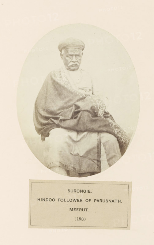 Surongie. Hindoo follower of parusnath. Meerut, Portrait of Shadi Seth, a man from the Jain Bania community (banker) from Bagpat, Uttar Pradesh, India, photograph, page, number: '(153)', inscription: 'SURONGIE., HINDOO FOLLOWER OF PARUSNATH., meerUT.', Bagpat, c. 1858 - in or before 1868, photographer, North India, paper, albumen print, height 129 mm x width 104 mm, head-gear: turban, banker, human races, peoples, nationalities, An oval sepia photograph of an older man seated and facing forward, wearing a rounded cap and a draped patterned shawl over lighter clothing, with his hands resting in his lap and a neutral facial expression; the portrait is mounted on a pale background with a rectangular label area beneath the image.
