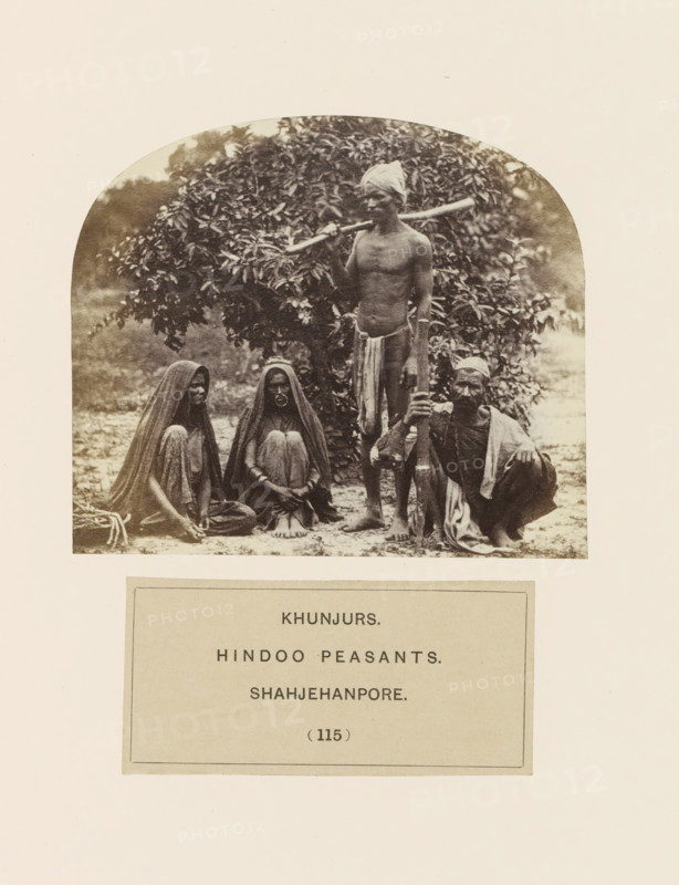 Four people from the Kanjar tribe of Shahjahanpur, Uttar Pradesh, India, photograph, page, number: '(115)', inscription, `: 'KHUNJURS., HINDOO PEASANTS., SHAHJEHANPORE', Shahjahanpur, c. 1858 - in or before 1868, photographer, North India, paper, albumen print, height 92 mm x width 107 mm, farmers, human races, peoples, nationalities, A sepia photograph framed by a rounded top shows four people posed outdoors in front of dense foliage; two seated women on the left wear long draped garments covering their heads and bodies and sit with knees drawn up, one of them displaying a large circular ornament on the face, a standing man in the center is bare-chested with a cloth wrapped around his head and a long piece of cloth tied around his waist while he rests a long pole across his shoulders, and a crouching man on the right wears a headwrap and loose garments and holds a tall staff upright with one hand.