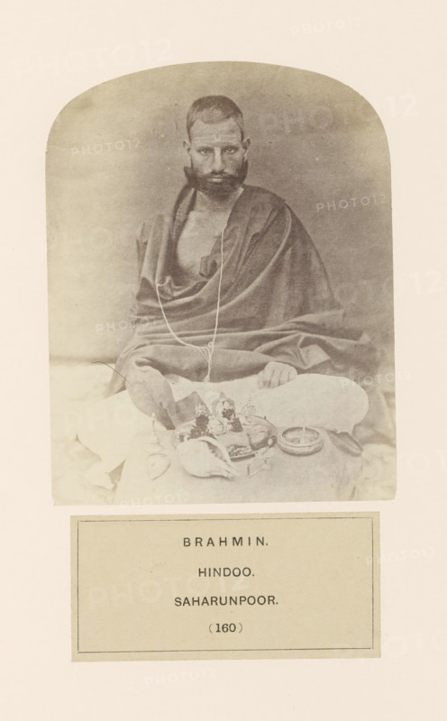Portrait of an unknown brahmin (Hindu priest) with items for his morning prayers, from Saharanpur, Uttar Pradesh, India, photograph, page, inscription: 'BRAHMIN., HINDOO., SAHARUNPOOR.', number: '(160)', Saharanpur, c. 1858 - in or before 1868, photographer, North India, paper, albumen print, height 123 mm x width 95 mm, human races, peoples, nationalities, ritual practices with particular objectives, Hinduism, Buddhism, Jainism, A sepia-toned photograph shows a seated man with short hair and a full beard wearing draped cloth over his shoulders and body, a thin cord hanging around his neck, his hands resting on his knees; in front of him are various ritual objects including a conch shell, small bowls, a plate with items arranged on it, and a shallow dish, all placed on a cloth surface, with a plain backdrop and rounded top framing the image.
