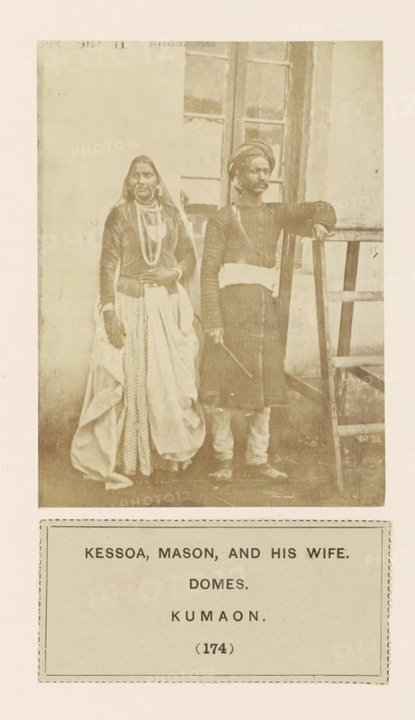 Portrait of a couple in front of a house in Kumaon, Uttarakhand, India, photograph, page, number: '(174)', inscription: 'KESSOA, MASON, AND HIS WIFE., DOMES., KUMAON.', Kumaon, c. 1859 - in or before 1869, photographer, paper, albumen print, height 113 mm x width 85 mm, couple of lovers, human races, peoples, nationalities, A faded sepia photograph shows a man and a woman standing side by side in front of a building with a paneled window; the woman on the left wears layered clothing including a long patterned skirt, a draped outer cloth, long sleeves, necklaces, and a head covering, while the man on the right wears a long tunic with a sash at the waist, a turban, leggings, and holds a stick in one hand with his other arm resting on a wooden ladder positioned beside him.