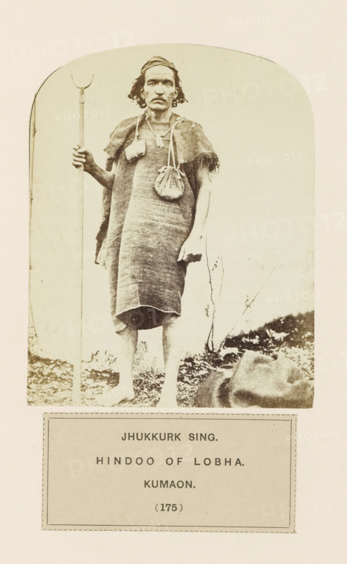 Portrait of Jhukkurk Sing, a man with a walking stick from Kumaon, Uttarakhand, India, photograph, page, inscription: 'JHUKKURK SING., HINDOO OF LOBHA., KUMAON.', number: '(175)', Kumaon, c. 1859 - in or before 1869, photographer, paper, albumen print, height 122 mm x width 96 mm, human races, peoples, nationalities, A sepia-toned photograph of a barefoot man standing on uneven ground, wearing a woven sleeveless garment that reaches to his knees and a small fringed shoulder covering, with a small pouch hanging from a strap across his chest and another pouch at his side; he holds a tall staff with a curved top in one hand, wears a headband, and has shoulder-length hair and a mustache, while a cloth or bundle lies on the ground near his feet and sparse vegetation appears behind him.