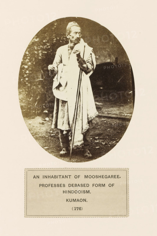 Portrait of an unknown man from the Munsyari region of Kumaon, Uttarakhand, India, photograph, page, inscription: 'AN INHABITANT OF MOOSHEGARHEE., PROFESSES DEBASED FORM OF, HINDOOISM., KUMAON.', number: '(176)', Munsyari, c. 1859 - in or before 1869, photographer, paper, albumen print, height 118 mm x width 95 mm, Hinduism, Buddhism, Jainism, human races, peoples, nationalities, A sepia-toned oval photograph shows an older man standing outdoors on a dirt surface, wearing layered traditional clothing including a long garment and a draped shawl, with a small cap on his head; he holds a long staff with one hand and a small object in the other, and his face shows a mustache and beard while he looks slightly to the side, with blurred foliage and dark background behind him.