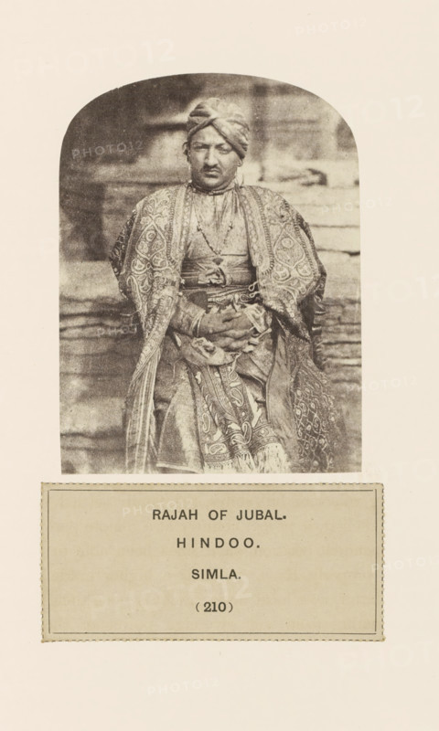 Portrait of the raja (king) of Jubbal near Shimla, Himachal Pradesh, India, photograph, page, number: '(210)', inscription: 'RAJAH OF JUBAL., HINDOO., SIMLA.', Shimla, c. 1859 - in or before 1869, photographer, paper, albumen print, height 100 mm x width 76 mm, ruler, sovereign, human races, peoples, nationalities, A sepia-toned portrait of a seated person wearing a patterned robe and a turban, hands clasped in their lap, shown inside a rounded-arch frame above a rectangular label panel.