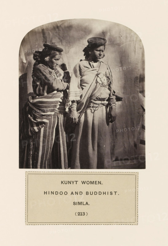 Two unknown women in traditional dresses from the Kanet community of Shimla, Himachal Pradesh, India, photograph, page, The Kanet community is also known as Kunait., inscription: 'KUNYT WOMEN., HINDOO AND BUDDHIST., SIMLA.', number: '(213)', Shimla, c. 1859 - in or before 1869, photographer, paper, albumen print, height 114 mm x width 95 mm, folk costume, regional costume, human races, peoples, nationalities, A black-and-white photograph showing two women standing side by side inside an arched frame with a plain backdrop, both wearing layered clothing and round caps; the woman on the left faces slightly toward the other, wearing a striped wrap and patterned garments with her hair tied back and one hand raised near her face, while the woman on the right faces forward with a long skirt and a sash draped across her chest, jewelry visible at her ears, and both hands hanging by her sides.