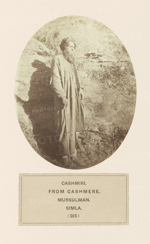 Portrait of an unknown merchant from Kashmir in Shimla, Himachal Pradesh, India, photograph, page, number: '(215)', inscription: 'CASHMIRI., FROM CASHMERE., MUSSULMAN., SIMLA.', Shimla, c. 1859 - in or before 1869, photographer, paper, albumen print, height 120 mm x width 95 mm, merchant, salesman, head-gear: turban, human races, peoples, nationalities, A sepia-toned oval photograph shows a bearded man standing against a rocky backdrop. He wears a long robe and a turban, and his hands rest near a walking staff. The rock surface behind him casts a shadow, and grasses or small plants are visible on the uneven ground. The image is mounted on a pale page with a rectangular label area below the oval.