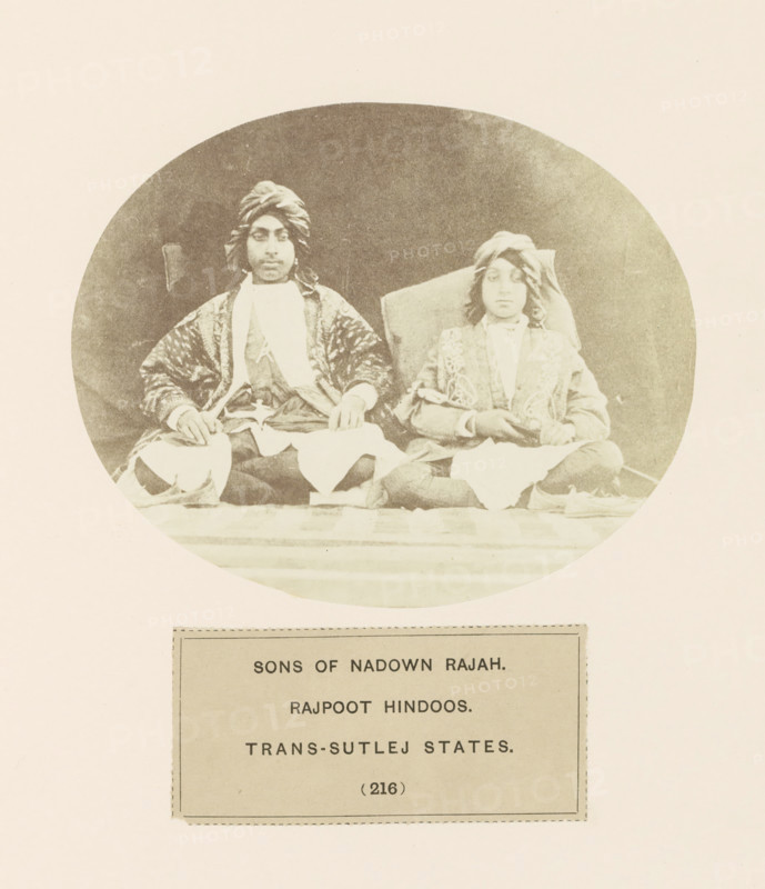 Portrait of two sons of the raja (king) of Nadown near Kangra, Himachal Pradesh, India, photograph, page, number: '(216)', inscription: 'SONS OF NADOWN RAJAH., RAJPOOT HINDOOS., TRANS-SUTLEJ STATES.', Kangra, c. 1859 - in or before 1869, photographer, paper, albumen print, height 104 mm x width 129 mm, head-gear: turban, human races, peoples, nationalities, A faded circular sepia photograph shows two seated individuals wearing patterned robes and turbans, positioned against a dark backdrop; both sit cross-legged on a surface with draped cloths over their laps, the figure on the left wearing a darker richly patterned outer garment and a light inner scarf while the figure on the right wears a lighter embroidered coat and has longer hair visible beneath the turban, both resting their hands on their knees, and below the photograph there is a rectangular label with printed lines and a border.