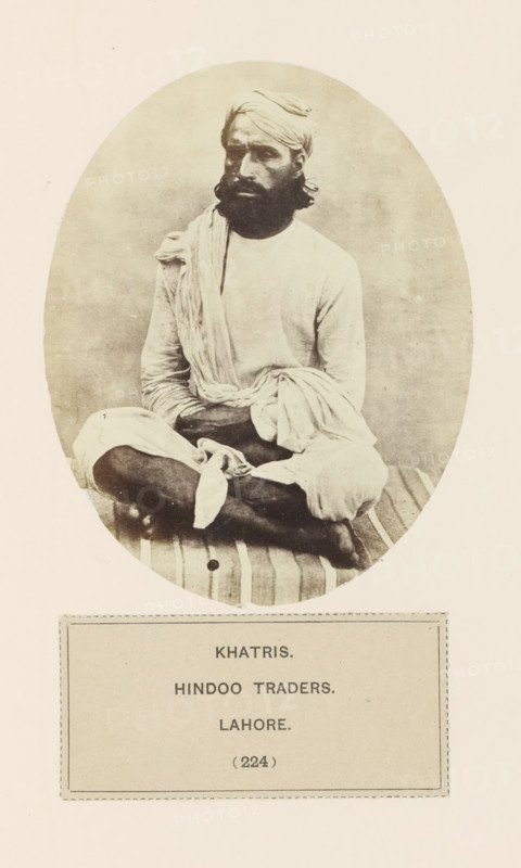 Portrait of an unknown trader from the Khatri community of Lahore, Punjab, Pakistan, photograph, page, inscription: 'KHATRIS., HINDOO TRADERS., LAHORE.', number: '(224)', Lahore, c. 1859 - in or before 1869, photographer, paper, albumen print, height 120 mm x width 95 mm, head-gear: turban, human races, peoples, nationalities, persons engaged in trade and commerce, A sepia-toned oval photograph of a seated man wearing a turban and loose light-colored clothing, with a long beard and mustache, sitting cross-legged on a striped cushion or mat, one arm resting on his knee and fabric draped over his shoulder and lap, set against a plain background and mounted on a pale page with a rectangular bordered label beneath the oval.