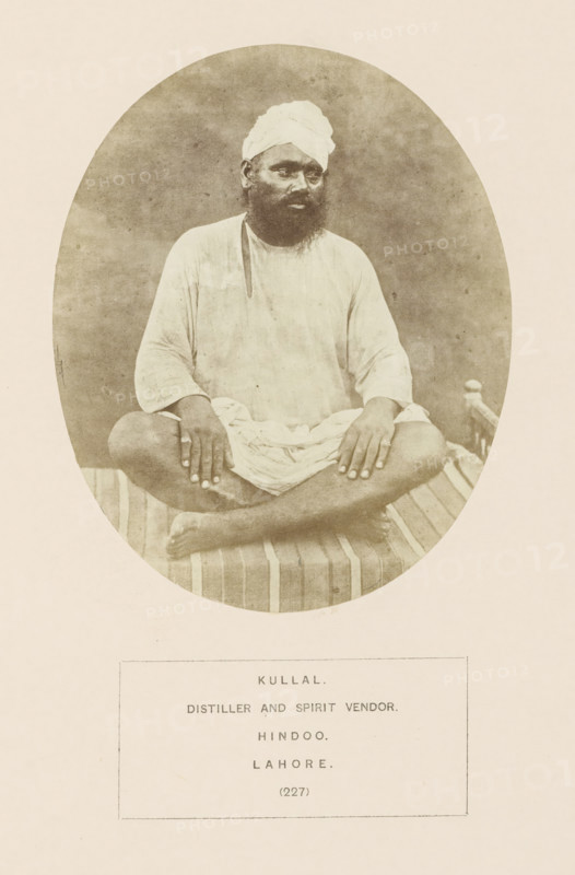 Portrait of a distiller and seller of liquor (kullal) from Lahore, Punjab, Pakistan, photograph, page, number: '(227)', inscription: 'KULLAL., DISTILLER AND SPIRIT VENDOR., HINDOO., LAHORE.', Lahore, c. 1862 - in or before 1872, photographer, paper, albumen print, height 129 mm x width 104 mm, distilled spirits, human races, peoples, nationalities, A sepia-toned oval photograph shows a seated man wearing a light-colored turban and loose long-sleeved garment, sitting cross-legged on a striped woven cot; his hands rest on his knees, he has a dark beard and mustache, and the plain backdrop is textured and neutral in tone.