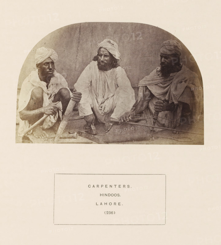 Portrait of three unknown carpenters with tools from Lahore, Punjab, Pakistan, photograph, page, inscription: 'CARPENTERS., HINDOOS., LAHORE.', number: '(236)', Lahore, c. 1862 - in or before 1872, photographer, paper, albumen print, height 89 mm x width 136 mm, carpenter, human races, peoples, nationalities, Three men sit close together on a mat against a plain backdrop, each wearing draped cloth garments and turbans; the man on the left is squatting with bare feet visible and holds a long wooden pole across his lap, the central man sits with legs bent and hands resting on the mat, looking slightly toward the left while his clothing hangs in loose folds, and the man on the right sits with a long beard and grasps a rounded wooden handle attached to a curved tool resting across his knees.