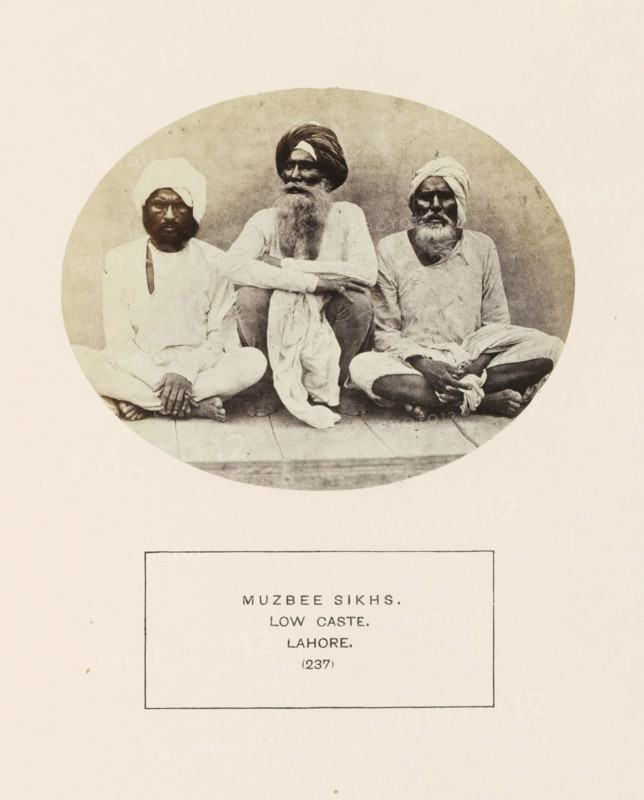 Portrait of three unknown men from the Mazhabi Sikh community of Lahore, Punjab, Pakistan, photograph, page, number: '(237)', inscription: 'MUZBEE SIKHS., LOW CASTE., LAHORE.', Lahore, c. 1862 - in or before 1872, photographer, paper, albumen print, height 95 mm x width 120 mm, human races, peoples, nationalities, Three seated men appear inside a circular frame, each wearing light-colored loose clothing and turbans, with visible facial hair and bare feet; the man in the center has a long beard and folded arms resting on his knees, the two men on either side sit cross-legged with their hands resting on their legs, and a simple floorboard surface is visible beneath them.