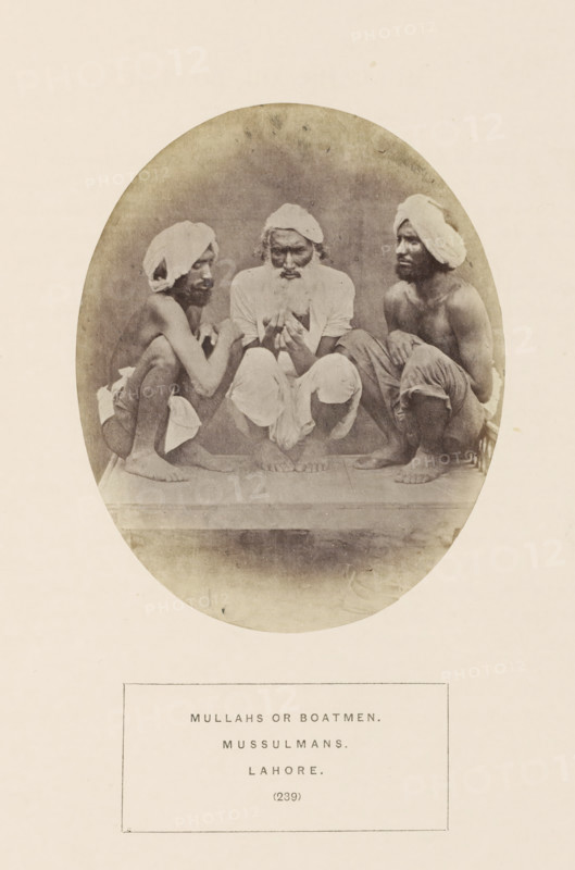 Portrait of three unknown bargemen from Lahore, Punjab, Pakistan, photograph, page, inscription: 'MULLAHS OR BOATMEN., MUSSULMANS., LAHORE.', number: '(239)', Lahore, c. 1862 - in or before 1872, photographer, paper, albumen print, height 128 mm x width 104 mm, human races, peoples, nationalities, A sepia-toned oval photograph shows three barefoot men seated close together on a raised surface, each wearing light cloths wrapped around their heads and simple lower garments; the central man has a full beard and is looking downward while holding something with both hands, the two men on either side are crouched facing inward with visible lean torsos and folded arms, and the background is plain and slightly mottled.