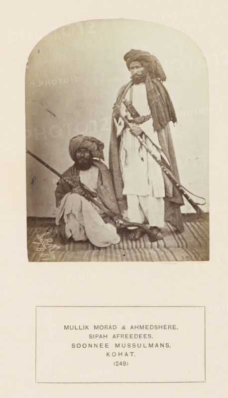Portrait of two men with muskets from the Sepah Afridi tribe of Kohat, Khyber Pakhtunkhwa, Pakistan, photograph, page, inscription: 'MULLIK MORAD & AHMEDSHERE., SIPAH AFREEDEES., SOONNEE MUSSULMANS., KOHAT.', number: '(249)', Kohat, 1861, photographer, paper, albumen print, height 115 mm x width 87 mm, firearms: rifle, human races, peoples, nationalities, A sepia-toned photograph shows two men wearing turbans and loose traditional clothing; one man is seated on a striped rug holding a long rifle across his lap, and the other man stands beside him holding a long firearm and wearing a draped shawl over his shoulders.