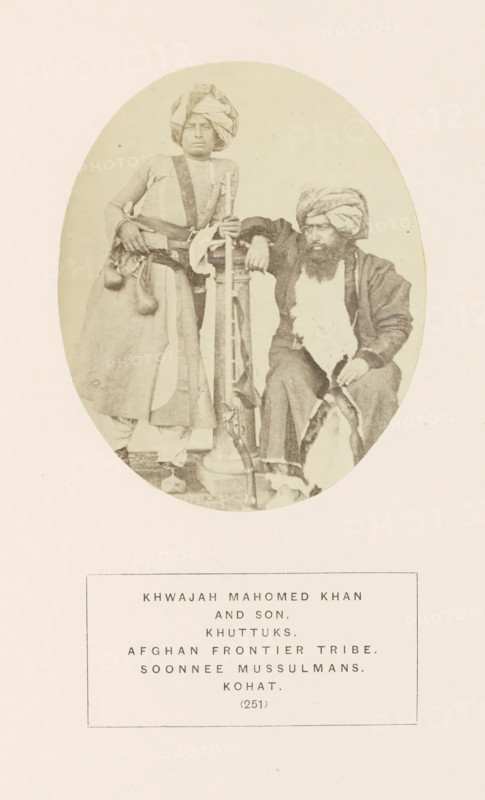 Portrait of Khwajah Mahomed Khan and his son from the Khattak tribe of Kohat, Khyber Pakhtunkhwa, Pakistan, photograph, page, number: '(251)', inscription: 'KHWAJAH MAHOMED KHAN, AND SON., KHUTTUKS., AFGHAN FRONTIER TRIBE., SOONNEE MUSSULMANS., KOHAT.', Kohat, c. 1862 - in or before 1872, photographer, paper, albumen print, height 107 mm x width 87 mm, firearms: rifle, human races, peoples, nationalities, An oval sepia photograph shows two men in traditional clothing against a plain background; the man on the left stands with a turban wrapped around his head, wearing a long garment with a sash and a belt from which small pouches hang, one hand resting on a tall vertical object and the other on his hip, while the man on the right sits with a turban and a full beard, wearing layered robes and holding a curved object across his lap, his elbow resting on the same vertical object between them.