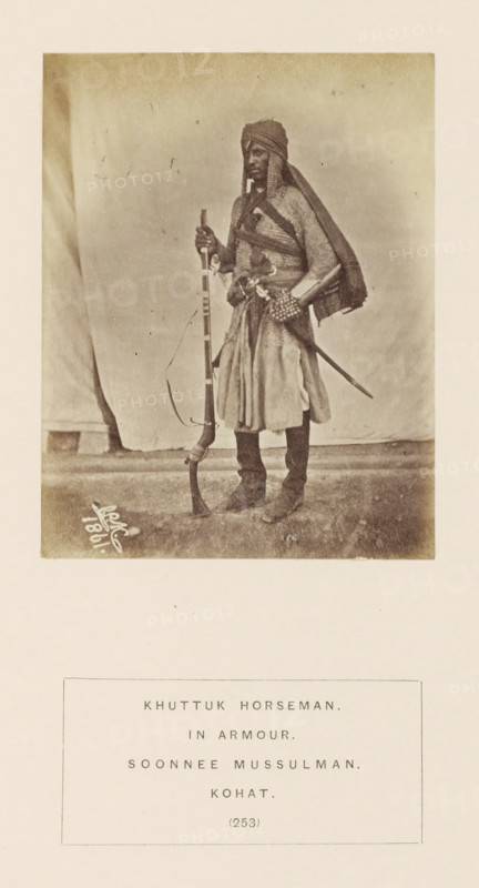 Portrait of a horseman with a musket and saber from the Khattak tribe of Kohat, Khyber Pakhtunkhwa, Pakistan, photograph, page, number: '(253)', inscription: 'KHUTTUK HORSEMAN., IN ARMOUR., SOONNEE MUSSULMAN., KOHAT.', Kohat, 1861, photographer, paper, albumen print, height 110 mm x width 86 mm, riding a horse, ass, or mule, rider, horseman, firearms: rifle, human races, peoples, nationalities, hacking and thrusting weapons (with NAME), A full-length sepia photograph of a person standing on a textured floor against a cloth backdrop, wearing layered garments including a draped head covering and a long tunic with a belt, holding a long firearm upright with one hand and resting the other hand near a sheathed sword at the waist, with boots visible and a strap or sash crossing the chest, and a small signature or marking near the lower left of the photo.