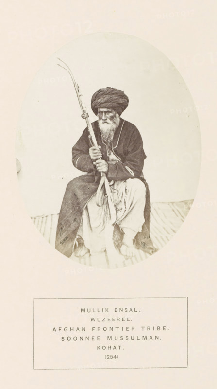 Portrait of Mullik Ensal, a man with a rifle from the Wazir tribe of Kohat, Khyber Pakhtunkhwa, Pakistan, photograph, page, The Wazir tribe is also known as Waziri., number: '(254)', inscription: 'MULLIK ENSAL., WUZEEREE., AFGHAN FRONTIER TRIBE., SOONNEE MUSSULMAN., KOHAT.', Kohat, c. 1862 - in or before 1872, photographer, paper, albumen print, height 120 mm x width 95 mm, head-gear: turban, firearms: rifle, human races, peoples, nationalities, An oval-framed sepia photograph shows an older man seated on a striped surface, wearing a dark turban and a dark long coat over lighter loose trousers, with a long white beard and a solemn facial expression, holding a long decorated rifle vertically across his body with both hands, his bare feet visible beneath his clothing.